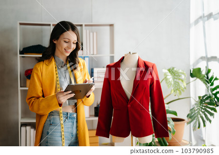 Fashion designer woman talking smart phone and using laptop with digital tablet computer in modern studio the clothes hanging on the racks . Fashion designer woman talking smart phone and using laptop with digital tablet computer in modern studio the clothes hanging on the racks . 103790985