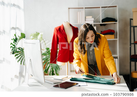 Caucasian young woman on desk in home office of fashion designer and holds tablet, laptop and smartphone. 103790986
