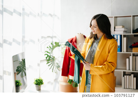 Caucasian young woman on desk in home office of fashion designer and holds tablet, laptop and smartphone. Caucasian young woman on desk in home office of fashion designer and holds tablet, laptop and smartphone. 103791127