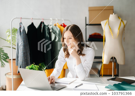 Caucasian young woman on desk in home office of fashion designer and holds tablet, laptop and smartphone. 103791366