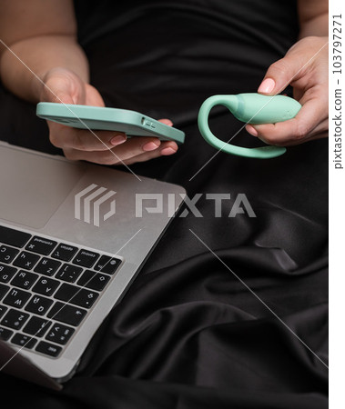 A woman lies on a bed and synchronizes the kegel machine with a smartphone. Girl watching video on laptop.  103797271