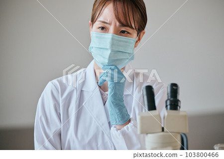A young woman in a white coat thinking with her hand on her chin at the desk A young woman in a white coat thinking with her hand on her chin at the desk 103800466