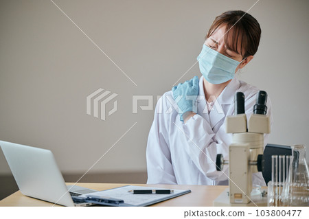 A young woman in a white coat holding her stiff shoulders with her hands 103800477