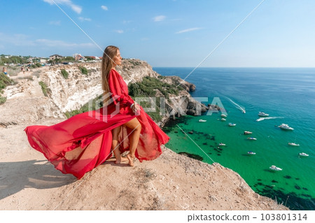Red Dress Woman sea Cliff. A beautiful woman in a red dress and white swimsuit poses on a cliff overlooking the sea on a sunny day. Boats and yachts dot the background. Red Dress Woman sea Cliff. A beautiful woman in a red dress and white swimsuit poses on a cliff overlooking the sea on a sunny day. Boats and yachts dot the background. 103801314