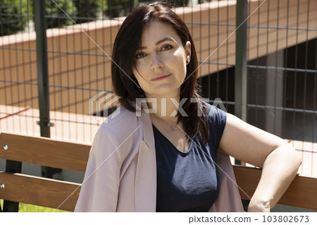Portrait Beautiful Brunette Mature Woman Sitting On A Bench In Residential Campus. Female Is Dressed In Casual Clothes. Rest And Relaxation. Horizontal Plane. Portrait Beautiful Brunette Mature Woman Sitting On A Bench In Residential Campus. Female Is Dressed In Casual Clothes. Rest And Relaxation. Horizontal Plane. 103802673