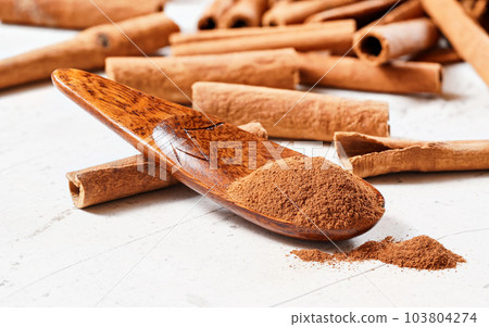 Grounded cinnamon powder in small wooden spice spoon on white board, blurred whole sticks in background, close up macro 103804274