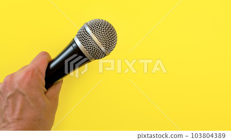 Man hand holds black and silver microphone over yellow background Man hand holds black and silver microphone over yellow background 103804389