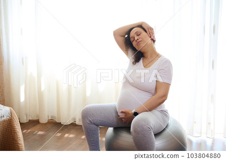 Portrait of multi ethnic pregnant cute woman, expectant gravid mother holding hand on her belly, doing stretching exercises, working out at home, sitting on a fitball. Pregnancy and maternity Portrait of multi ethnic pregnant cute woman, expectant gravid mother holding hand on her belly, doing stretching exercises, working out at home, sitting on a fitball. Pregnancy and maternity 103804880