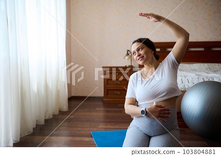 Delightful gorgeous pregnant woman holds her hand on her big bell, stretching her arm right, sitting in hero pose on a yoga mat, practicing prenatal fitness for wellness and healthy in pregnancy time 103804881