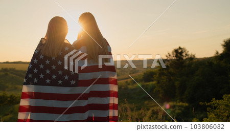 A mother with a teenage daughter with an American flag on their shoulders look at the sunset over a picturesque valley 103806082