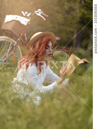Young redhead woman in dress and hat with retro bike at city park reading the book. Vacation Young redhead woman in dress and hat with retro bike at city park reading the book. Vacation 103806868