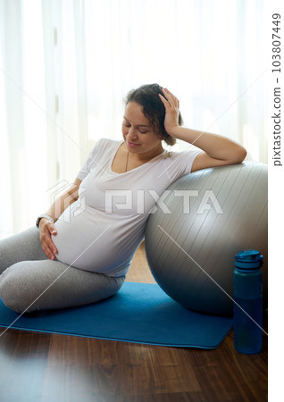 Happy pregnant woman gently stroking her belly, relaxing on a yoga mat after prenatal stretching exercises on fitball, smiling enjoying a healthy active lifestyle in pregnancy time and maternity leave 103807449