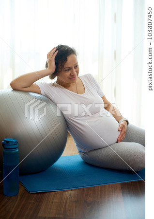 Gravid woman holding her hand on her pregnant belly, relaxing on exercise mat after prenatal yoga practice and meditation, expressing positive emotions of a healthy active lifestyle in pregnancy time 103807450