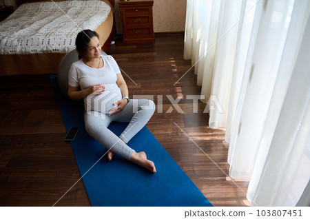 Top view of delightful relaxed pregnant woman in white t-shirt, sitting barefoot on a blue yoga mat, stroking her big belly, relaxing after prenatal relaxation exercises at home. Healthy pregnancy 103807451