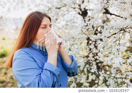 Sneezing young redhead woman with nose wiper among blooming trees in park. Portrait of sick women sneezes in white tissue, suffers from rhinitis and running nose. Symptoms of cold or allergy. 103810547