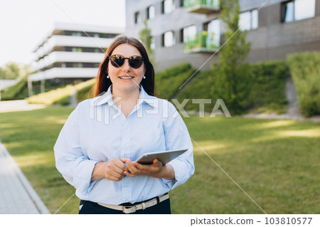 Young cheerful woman using digital tablet near modern building. Businesswoman wearing eyeglasses working online and standing on street. Successful business. Happy female using modern technology Young cheerful woman using digital tablet near modern building. Businesswoman wearing eyeglasses working online and standing on street. Successful business. Happy female using modern technology 103810577