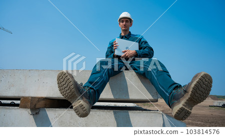 Caucasian male builder in hardhat sits on floor slabs and uses laptop at construction site. Caucasian male builder in hardhat sits on floor slabs and uses laptop at construction site. 103814576