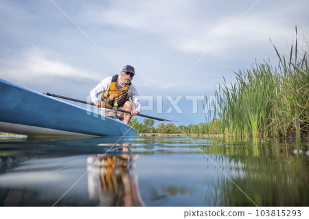 senior male paddler is paddling a stand up paddleboard on a calm lake in early spring, frog perspective from an action camera at water level 103815293