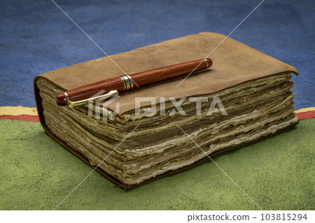 retro leather-bound journal with decked edge handmade paper pages and a stylish pen against abstract paper landscape, journaling concept 103815294