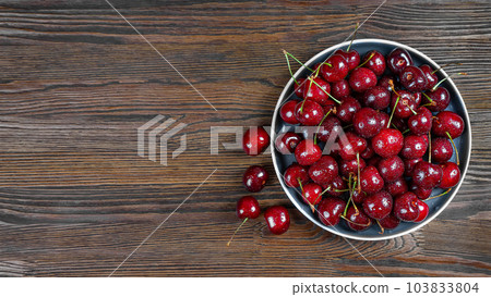 Fresh red ripe sweet cherry with water drops on plate on brown wooden background. Fresh red ripe sweet cherry with water drops on plate on brown wooden background. 103833804