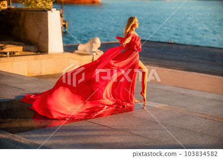 Sunrise red dress. A woman in a long red dress against the backdrop of sunrise, bright golden light of the sun's rays. The concept of femininity, harmony. 103834582