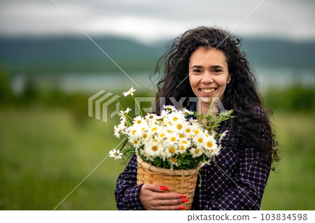 A woman stands on a green field and holds a basket with a large bouquet of daisies in her hands. In the background are mountains and a lake. A woman stands on a green field and holds a basket with a large bouquet of daisies in her hands. In the background are mountains and a lake. 103834598