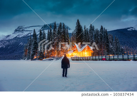 Traveler man standing in snowfield with wooden lodge glowing and pine forest in rocky mountains at Yoho national park 103837434