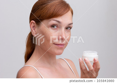 Portrait of cropped caucasian middle aged woman face with freckles holding opened jar with skin cream in hand on white background looking at camera 103841660