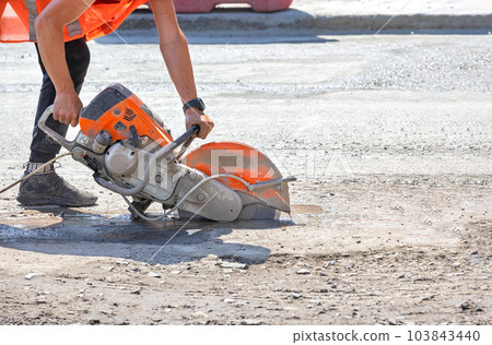 A petrol disc cutter in the hands of a worker cuts the asphalt concrete road surface on a sunny summer day. 103843440