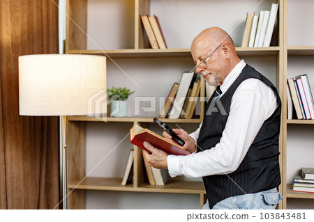 Senior man standing near bookshelves and reading book at home. Man using a magnifying glass. Man wearing white shirt and black waistcoat. Senior man standing near bookshelves and reading book at home. Man using a magnifying glass. Man wearing white shirt and black waistcoat. 103843481