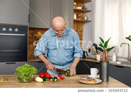 Retired senior man cooking from green salat, pepper and tomatoes. Man standing in kitchen interior. Man wearing jeans and shirt. 103843532
