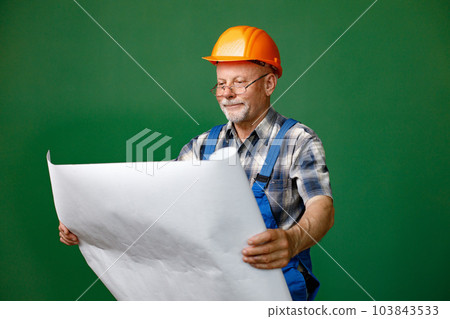 Studio portrait of a man isolated on green background. Seniour man worker in a blue plaid shirt and blue overall looking at scheme. Man wearing eyeglasses and orange helmet. 103843533
