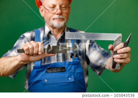 Studio portrait of a man isolated on green background. Seniour man worker in a blue plaid shirt and blue overall holds a calipers. Man wearing eyeglasses and orange helmet. 103843536