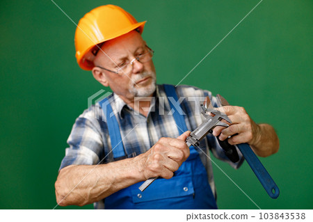 Studio portrait of a man isolated on green background. Seniour man worker in a blue plaid shirt and blue overall holds a straight pipe wrench or spanner. Man wearing eyeglasses and orange helmet. 103843538