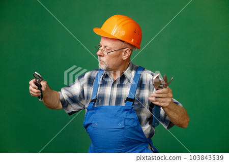 Studio portrait of a man isolated on green background. Seniour man worker in a blue plaid shirt and blue overall holds a straight pipe wrench or spanner. Man wearing eyeglasses and orange helmet. 103843539
