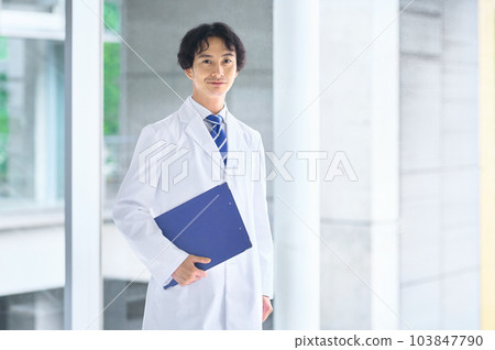 A young male doctor in a lab coat standing in the hospital corridor A young male doctor in a lab coat standing in the hospital corridor 103847790