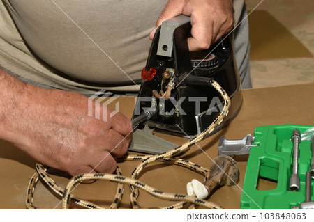 A man with a screwdriver unscrews the bolt connecting the electric cable with the iron. A man with a screwdriver unscrews the bolt connecting the electric cable with the iron. 103848063