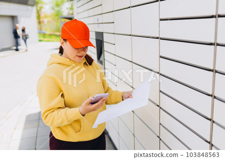 Young woman in red hat picks up parcel from automatic post office machine, Courier standing with phone and small box. Concept of fast delivery to automatic self lockers 103848563