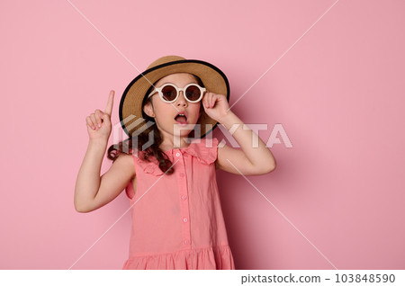 Amazed adorable child girl wearing sunglasses, pink dress and straw hat, pointing forefinger aside a copy space for your advertising text, on isolated pink background. Summer camp. Holidays concept 103848590