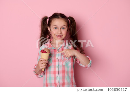 Adorable happy little child girl with two ponytails, wearing checkered shirt, smiling looking at camera, showing a waffle cone with delicious ice cream over isolated pink background. Food. Summer 103850517