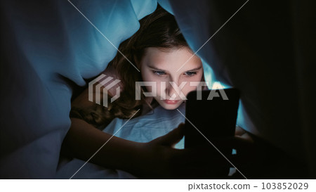 Screen time. Child insomnia. Late night chat. Internet addiction. Sleepless girl texting communicating on smartphone hiding under blanket in bed at home. 103852029