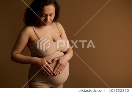 Beautiful multi ethnic adult pregnant woman, in beige underwear, holding hands and forming heart on her tummy isolated on beige background. Body positivity. Pregnancy. Childbearing. Nudity. Copy space 103852220