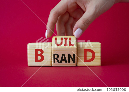 Build your brand symbol. Businessman hand points at wooden cubes with words Build and Brand. Beautiful red background. Build your brand and business concept. Copy space 103853008