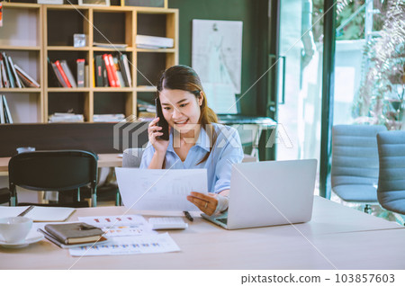 Business asian woman sitting at her desk with laptop using mobile phone chatting with customers or giving financial advice. Business asian woman sitting at her desk with laptop using mobile phone chatting with customers or giving financial advice. 103857603