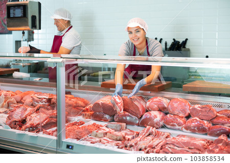 Young female seller shows demonstrates raw beef ribs in glass showcase of refrigerator. Young female seller shows demonstrates raw beef ribs in glass showcase of refrigerator. 103858534