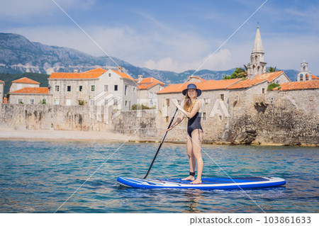 Young women Having Fun Stand Up Paddling in blue water sea in Montenegro. Against the backdrop of the Old Town of Budva. SUP 103861633