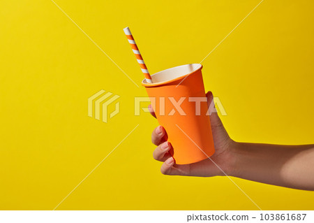 Against the light yellow background, female hand is holding a disposable orange paper cup with a paper straw. Scene for advertising drinking product. Copy space, summer concept Against the light yellow background, female hand is holding a disposable orange paper cup with a paper straw. Scene for advertising drinking product. Copy space, summer concept 103861687