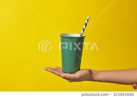 A green disposable paper cup with a paper straw placed in a woman's hand on a yellow background. Minimal scene for advertising and branding drinking product. Copy space A green disposable paper cup with a paper straw placed in a woman's hand on a yellow background. Minimal scene for advertising and branding drinking product. Copy space 103861688