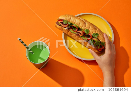 A bread filled meat, rolls and pate served with vegetable on round yellow plate and a disposable green paper cup decorated on orange background. Female hand is touching and prepare for eat 103861694