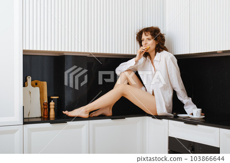Slender barefoot woman bite croissant while sitting on kitchen tabletop 103866644